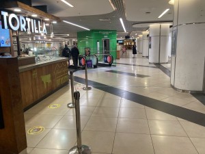 From second floor inside shopping center, facing  venue. Access via opening with queueing divider along right. Sidewalk to its right.