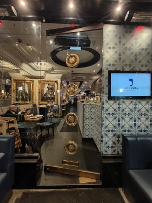 Facing glass entrance door. Golden metal circles engraved in centre of door. Push golden metal handle on left to open.