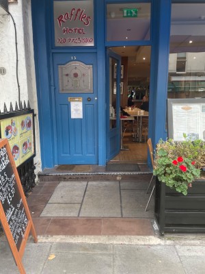 From street, facing venue. Planter on right. Glass doorway with blue wooden frame at back. Push silver handle on right to open.