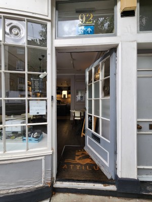 Single grey door with white doorframe. Doorbell on doorframe to the left at standing-height. Handle on left. Push to open.