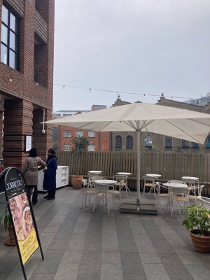 From sidewalk facing venue. Opening ahead. A-framed sign on left. Planters on right.
