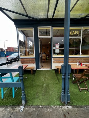 Opening ahead to before outdoor seating on left and right. Blue pillar on right. Path in centre to ramp then entrance at back.