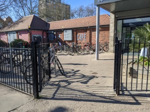 From pavement facing venue. Iron gate ahead. Push from centre to open.