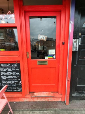 Bright red framed glass door. Push golden handle on left to open. Doorbell for assistance on right.