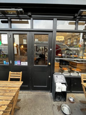Black framed glass door. Push golden handle on right to open. Small bump on threshold of door followed by doormat.