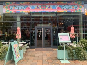 From street looking at venue front. Path to main entrance ahead. Tables and chairs both side of doors behind planters. Display left and right of path