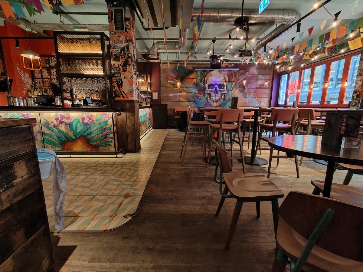 From welcome desk facing right. Path ahead going left. Bar at back left. Stools and high tables spread across right. Half booths at far back.