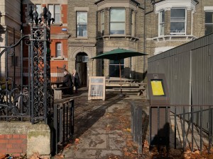 From sidewalk facing straight. Pathway through open gate. Outside bench seating area straight ahead. Archway on left front of building to inside area.