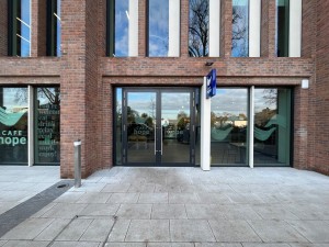 Glass doors ahead with metal handles in center and black frame. Push “Press to open” button on metal post left of doors.