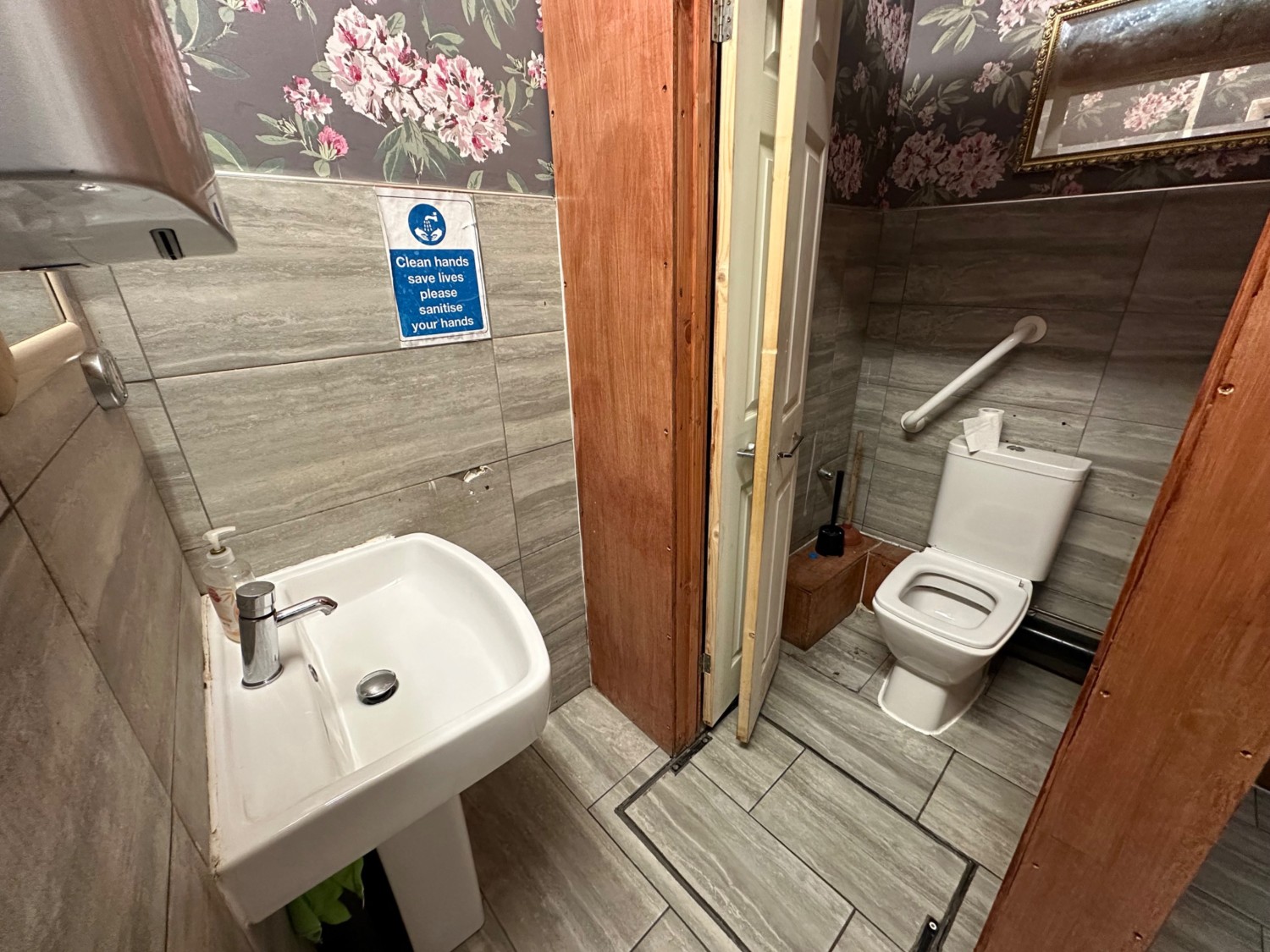 Facing slightly left, sink on left wall, bathroom cubicle on right. Toilet with single grab rail on back wall.