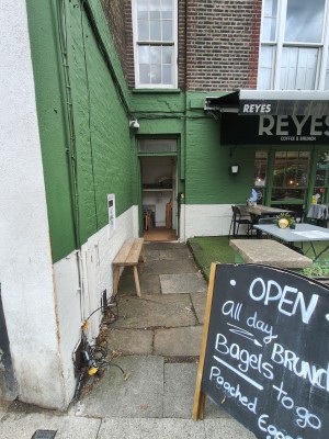 From street facing venue. A-frame sign on right. Path in centre to entrance at back. Bench before entrance on left.
