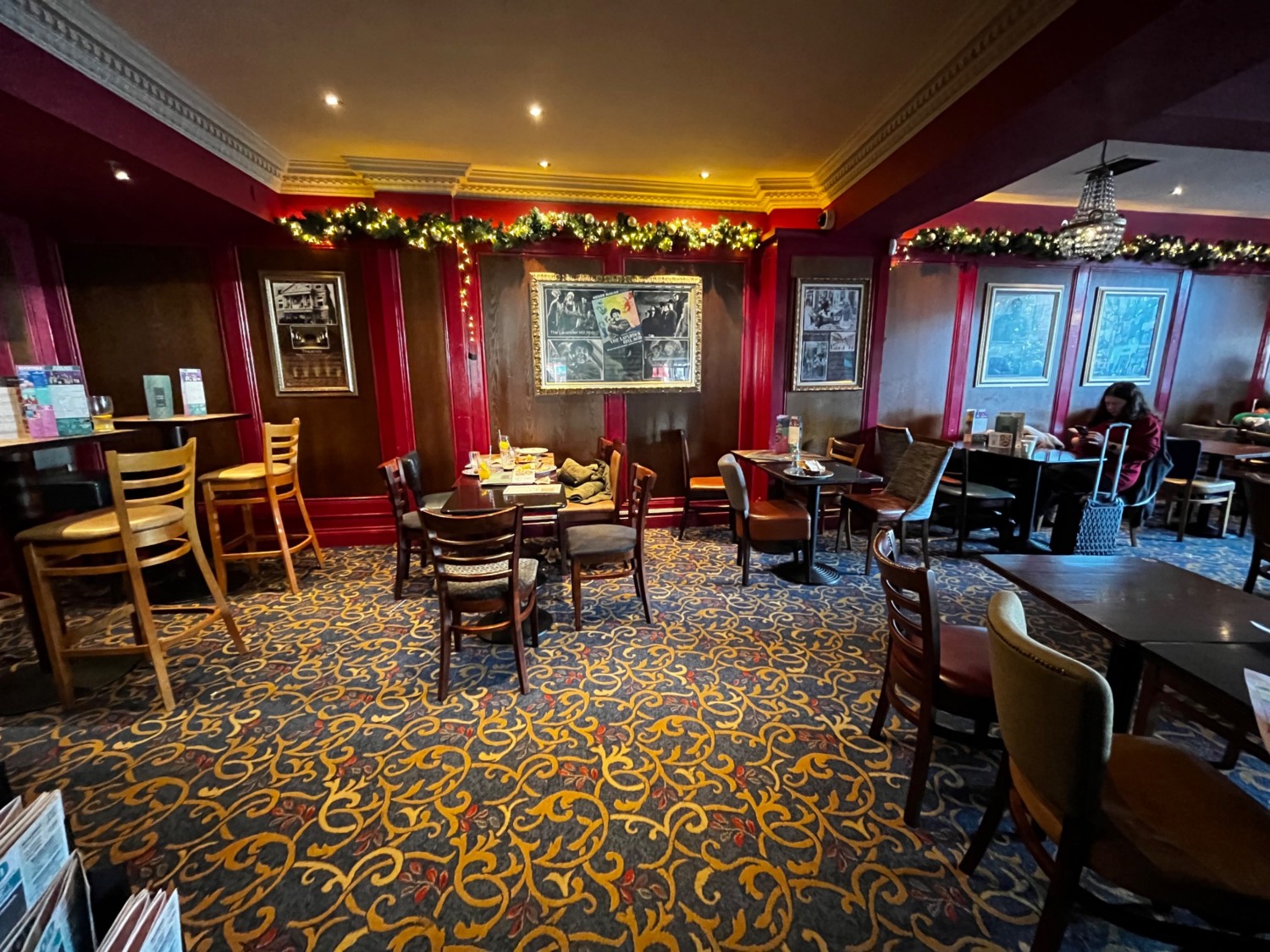 From archway facing straight. High tables and stools along left. Tables and chairs along back and spread on right.