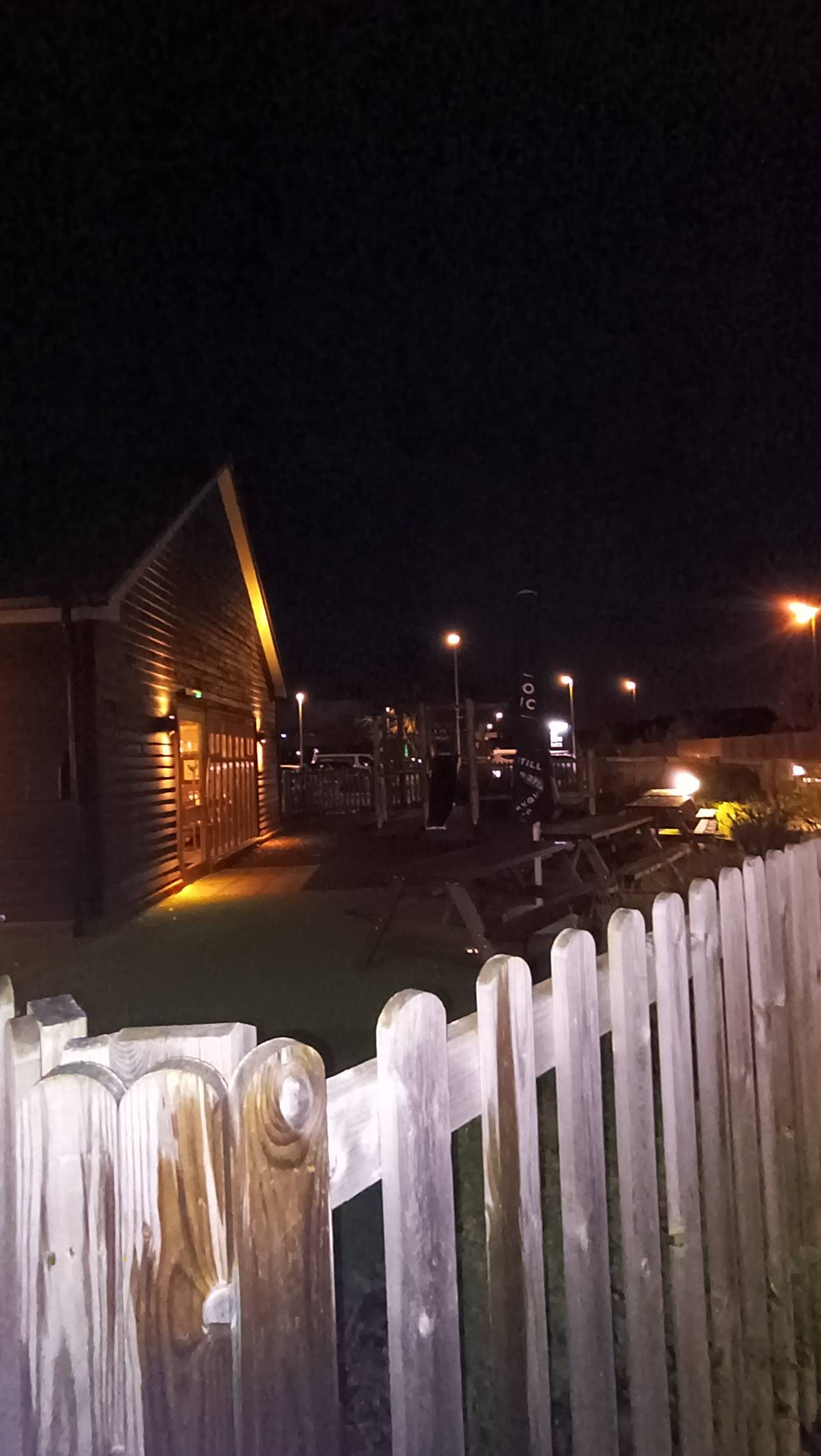 Children's wooden play area with climbing and slide. Picnic benches, not accessible for wheelchairs. Warm outside lighting. Small flower beds to right