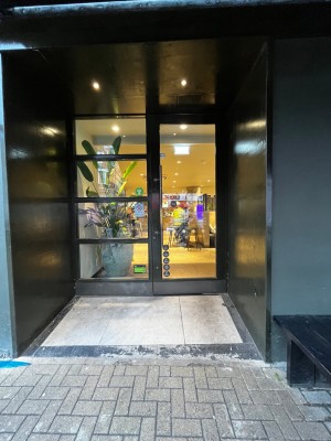 Open archway ahead to glass door with black frame on back. Pull door to open via handle on left. Ramp up from archway to door.