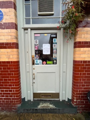 First door to entrance. White frames glass door with metal handle to left. Push to open.