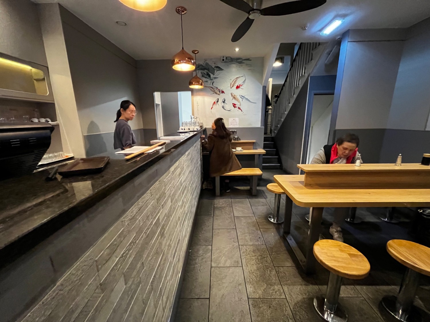From counter, facing straight. Counter along left. Table and benches at back. Tables and stools along right. Path to bathroom behind.