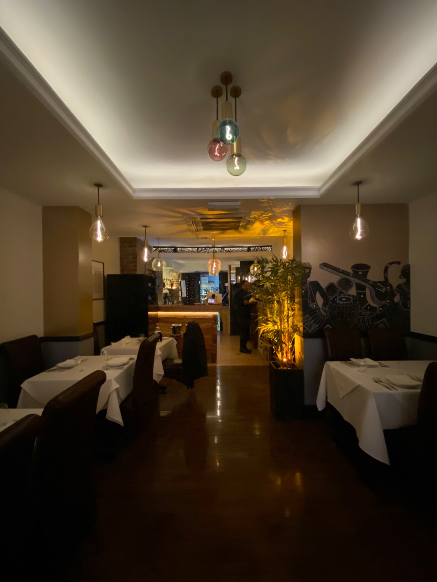 From middle of pathway, looking ahead. More tables and chairs along left and right walls. Bar counter on back left wall. Men’s bathroom on far right.