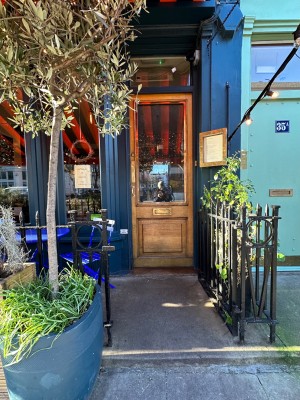 From street facing venue. Potted tree to direct left, railing right. Small bump before path to door. Wooden framed glass door. Push from left to open.