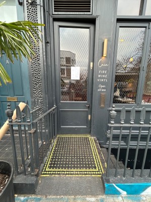Ramp leading up to door. Grey and glass panelled door. Push black handle from right to open.