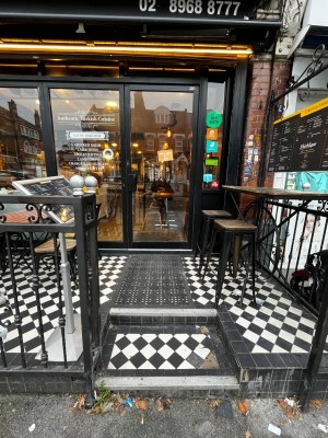 From pavement facing venue. Steps up to entrance ahead. Outdoor seating on right. Glass door with black frame at back. Push handle on left to open.