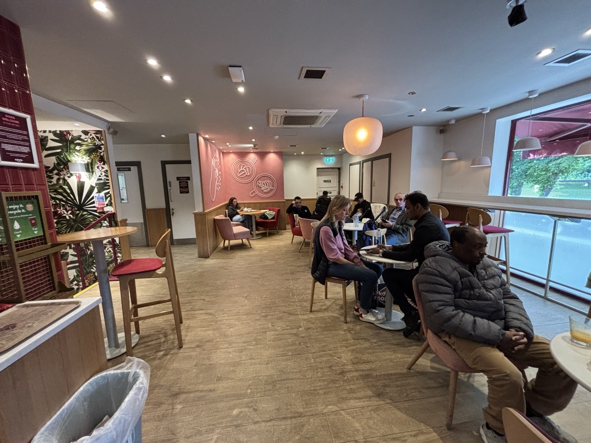 From opening facing left. Bin on left, high table behind. Tables and chairs along right. High table and stools along right wall.