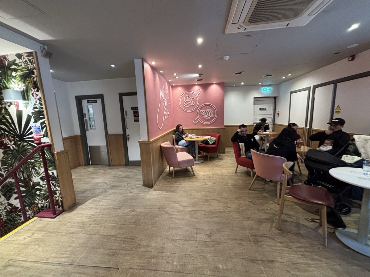 From bin facing straight. Stairs on left, opening to  bathroom behind. Tables with chairs and armchairs along right and on back.