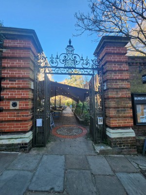From sidewalk facing venue. Metal gates ahead. Push from center to open. Path forward ahead after gate with outdoors seating along both sides.