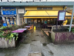 From street looking to venue. Gap in wooden fence followed by path straight to entrance. Seating along left of path.