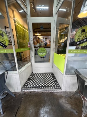 From street, facing venue. Opening ahead. Glass door with grey wooden door ahead. Pull silver handle on left to open.