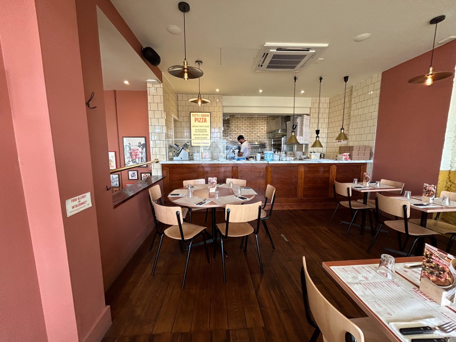 From archway, looking ahead. Tables and chairs directly ahead and along right wall. Pathway slightly along right. Open kitchen along back wall.