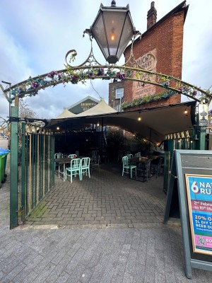 From street facing venue, gate archway ahead followed by outdoor area with seating on both sides. Path in middle to doorway.