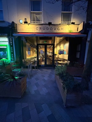 From pavement facing venue. Opening ahead. Planters to left and right of it. Outdoor seating behind. Path leads to doors.