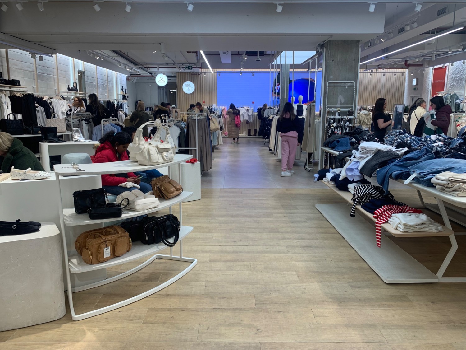 From after self checkout facing right. Product shelves along left. Path in centre to fitting room at back left. Product shelf on right.
