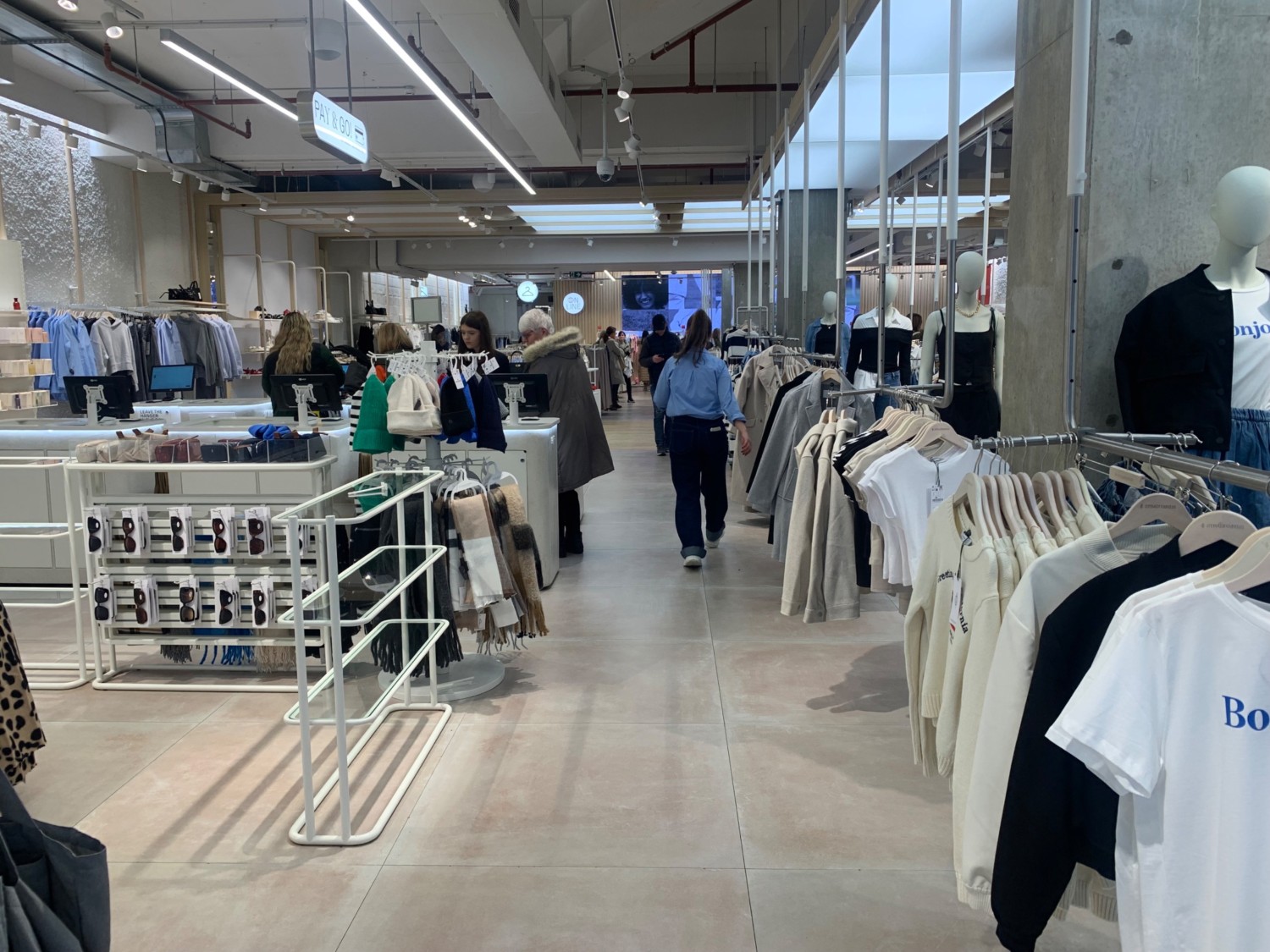 From end of clothing racks facing right. Racks along right. Path ahead to fitting rooms on back left. Product shelves on left. Self checkout behind.
