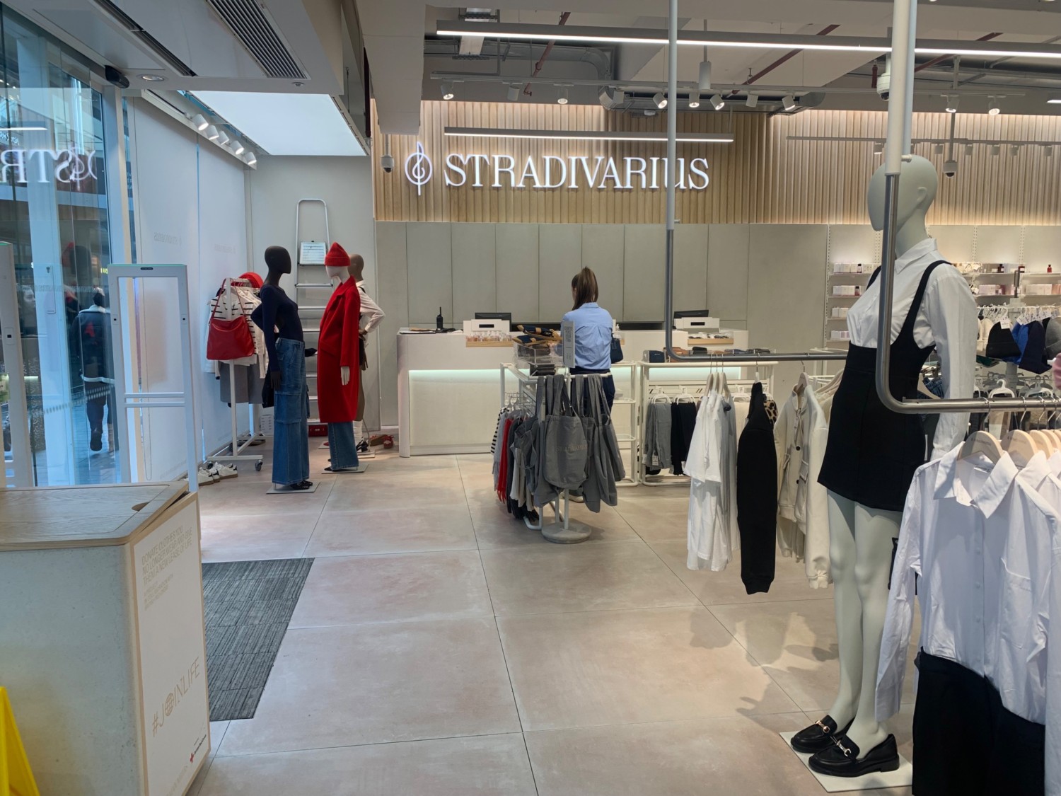 From entrance facing left. Donation box on left. Second entrance behind. Clothing racks along right side. Path in centre to service counter at back.