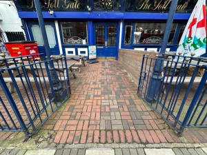 From street, facing venue. Blue railing gates ahead. Push from centre to open. Path behind continues straight ahead.
