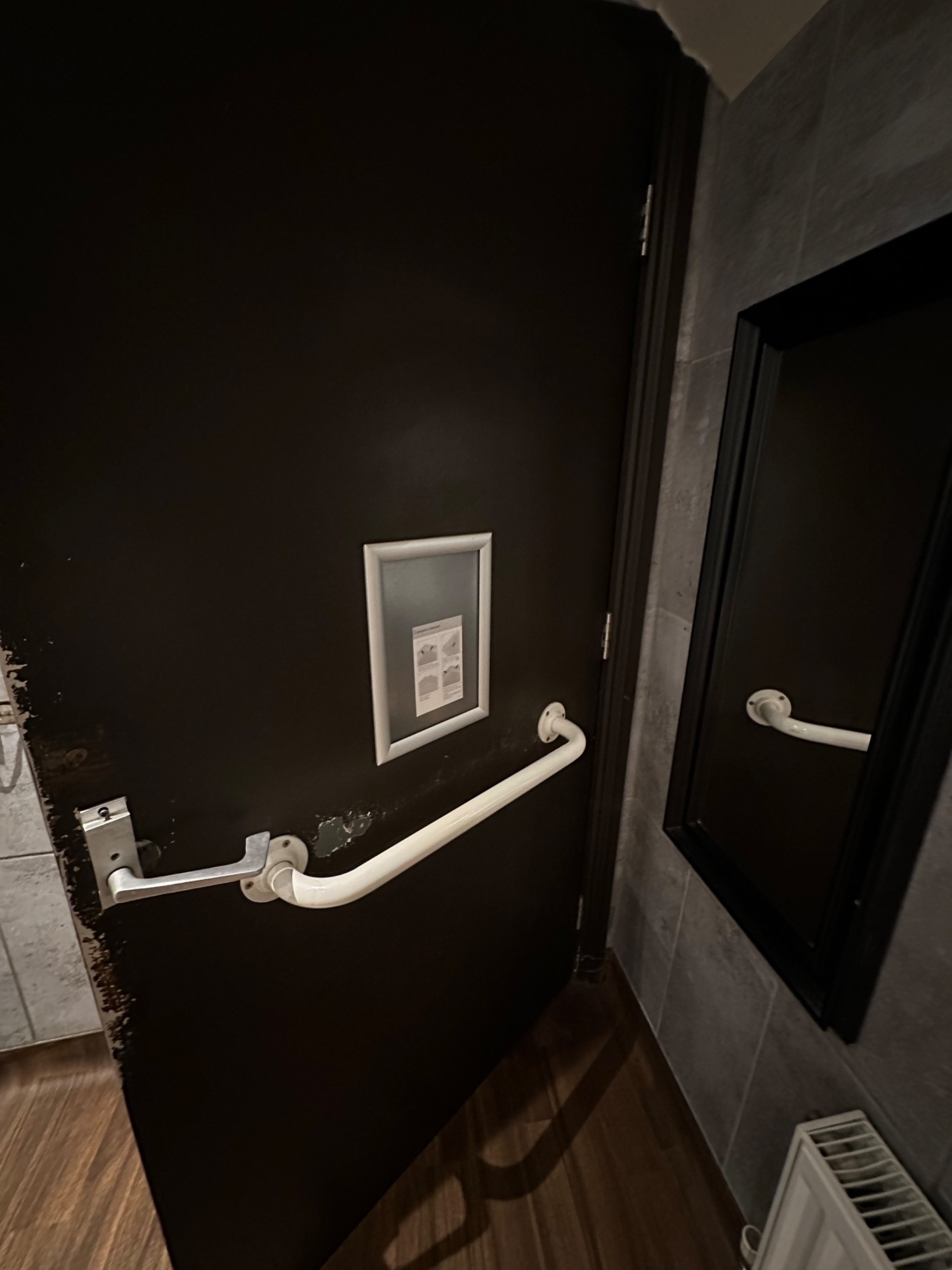 Back of door. White grab rail in centre. Push silver handle from left to open.