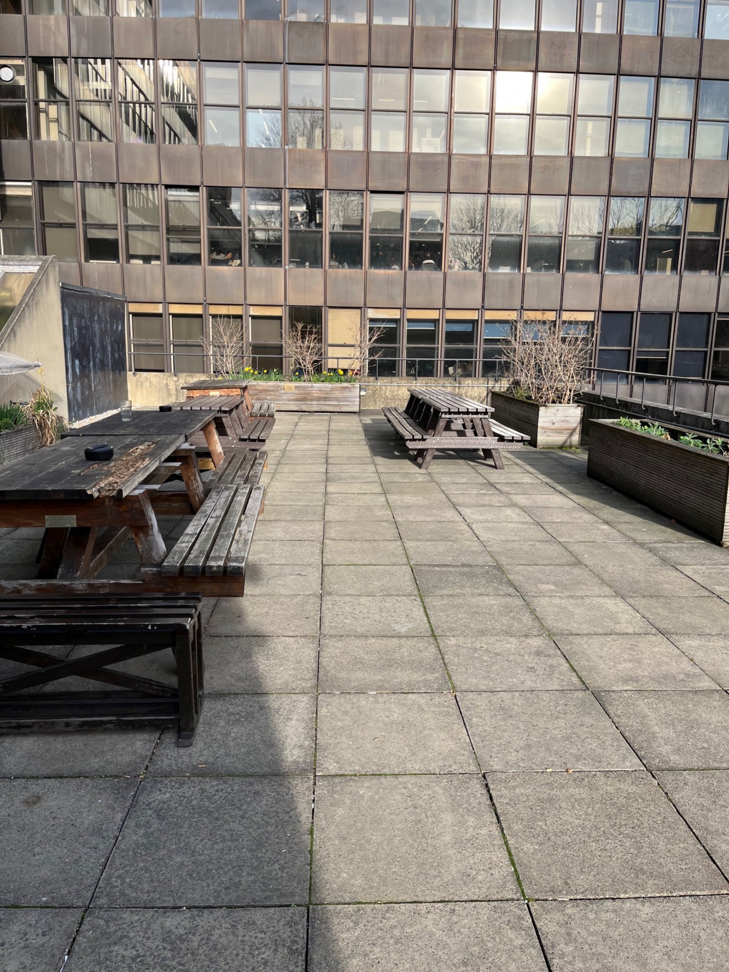 Open terrace with benches. Wide space. Benches are attached to table tops. Floor is grey and benches are brown