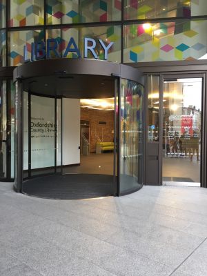 Photograph of the entrance to Oxfordshire County Library, showing the large automatic double doors on the left, and the manual, single door on the right. All doors are made of glass, and the