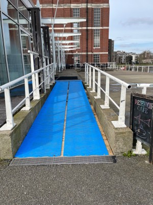 From left of venue facing right. Ramp up to main entrance ahead. White handrails to left and right of ramp.