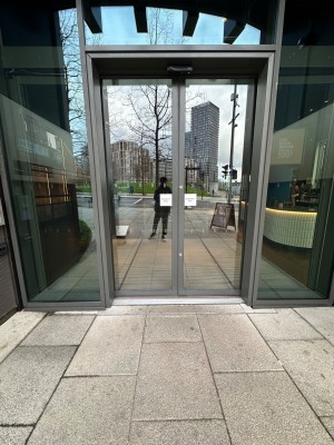 Double automatic glass doors within grey frame from street leading to downstairs entrance area.