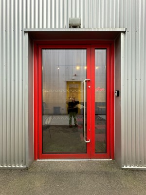 Glass panelled door with red frame. Silver handle towards right of door. Push to open.
