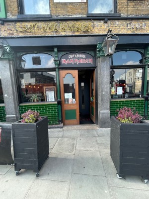 From street, facing venue. Opening ahead between planters on left and right. Path down middle leads to entrance on back wall.