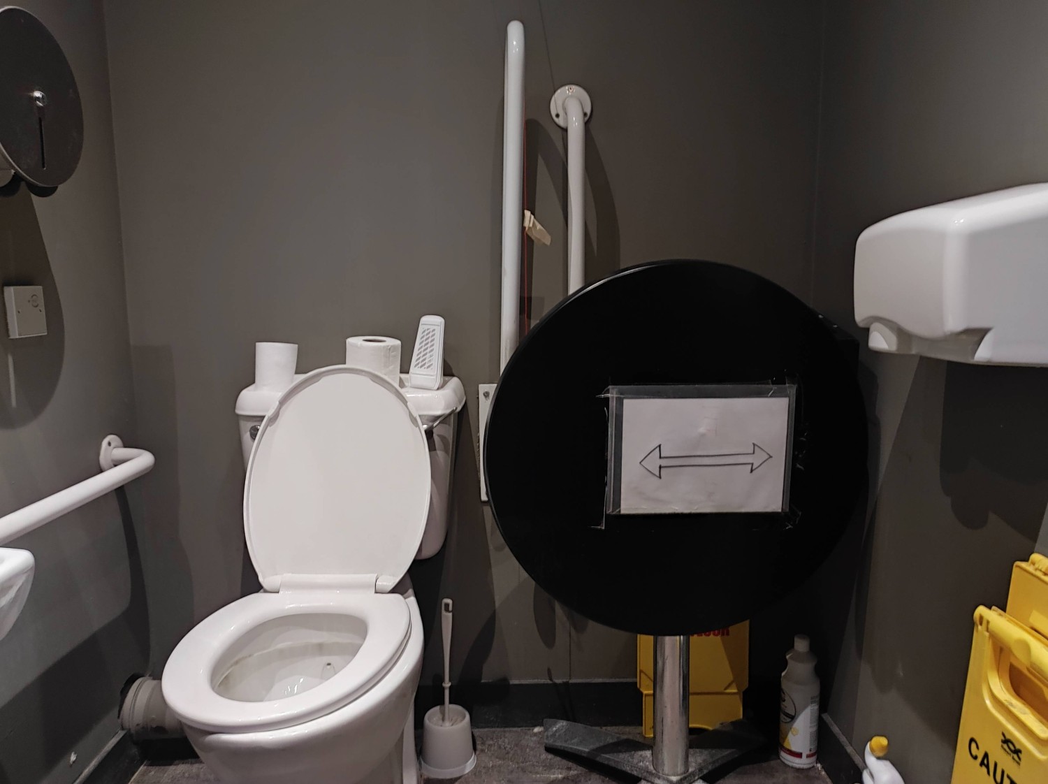 Wider image if the previous including a standing high hand dryer and the table blocking a transfer space with cleaning products blocking the floor