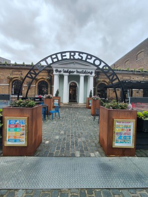 From street, facing venue. Opening ahead with planters on both sides. Outdoor seating after. Path ahead to doorway at back.