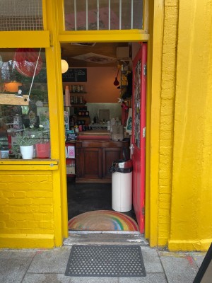 From sidewalk facing entrance. Red framed door with yellow doorframe ahead. Handle on left. Push to open inwards.