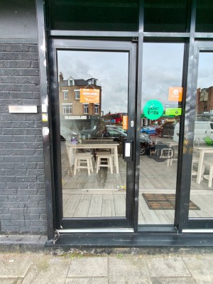From sidewalk facing entrance. Black framed single glass door ahead. Push handle on right to open inwards.