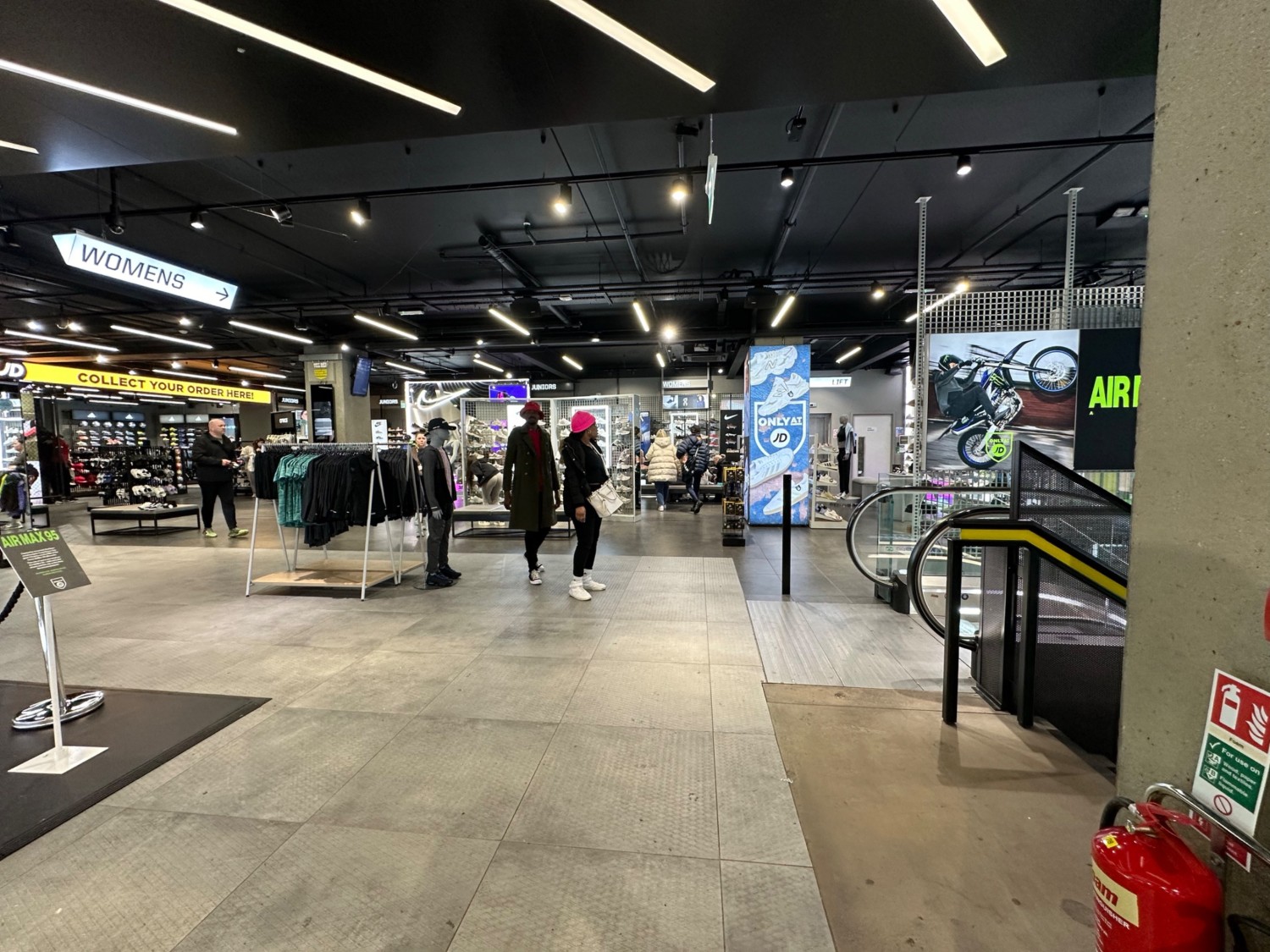 From top of stairs and escalator facing right. Display area on left. Stairs and escalator doing downstairs on right. Path ahead leads to more displays