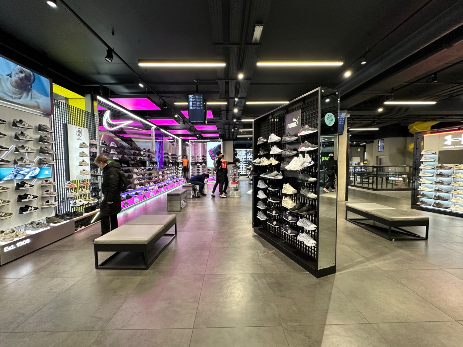From after first pillar facing right. Central shoe display ahead and shoe wall along left wall. Benches on left and right of shoe display ahead.