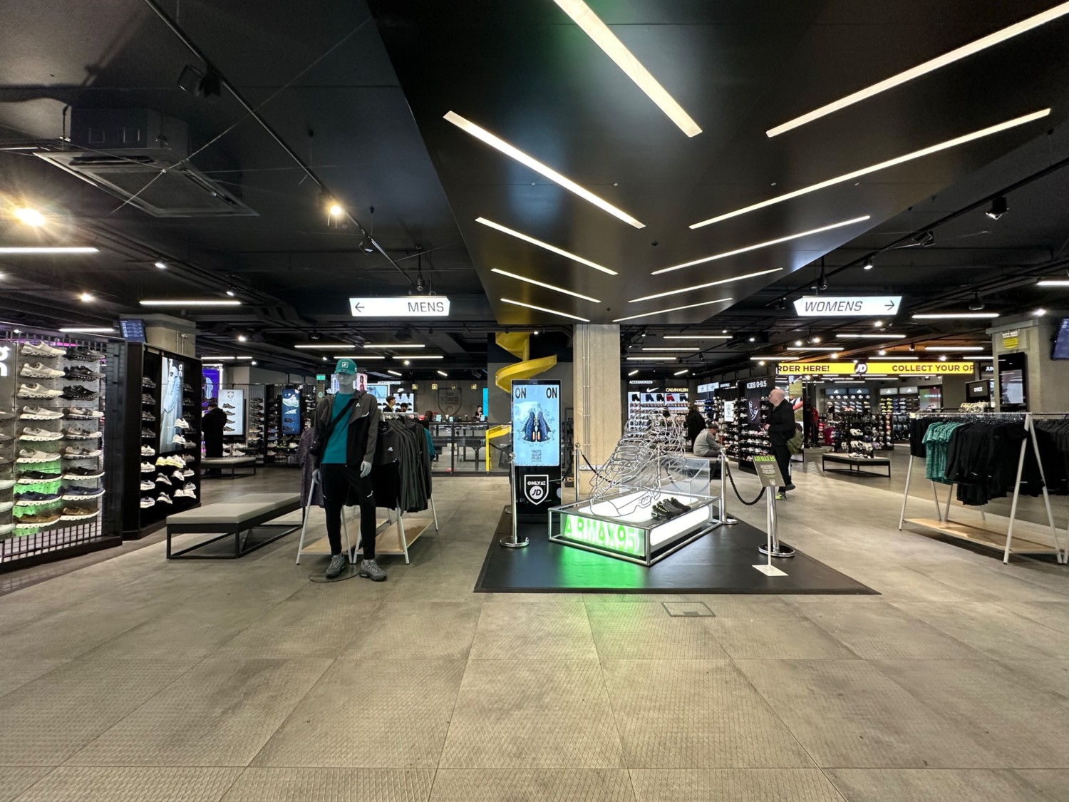 From top of stairs and escalator facing straight. Display area ahead obstructing path. Clothes rack to right. Shoe rack and bench to left.